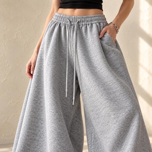 May include: Light grey sweatpants featuring a high-waisted design with an elastic waistband and a matching drawstring. These wide-leg trousers are crafted from a soft, heathered fabric and include side pockets for functional storage.