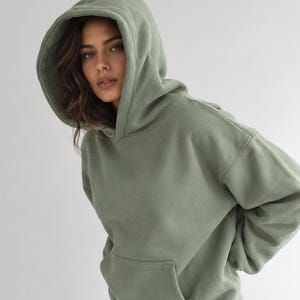 May include: A sage green hooded sweatshirt featuring a large, deep hood, long sleeves with ribbed cuffs, and a front kangaroo pocket. The garment is constructed from a solid-colored fabric and is paired with blue denim jeans.