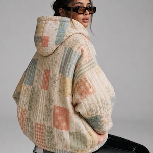 Peut inclure: Une veste à capuche matelassée présentant un motif patchwork avec divers imprimés floraux, rayés et à carreaux dans des tons sourds de crème, vert sauge, rose poudré et bleu clair. La veste a des manches longues et une coupe ample.