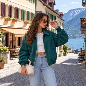 May include: A dark green zip-up jacket with a collar and long sleeves, worn over a white crop top and light blue denim jeans. The outfit is accessorized with a small beige handbag, gold layered necklaces, and thin-framed sunglasses.