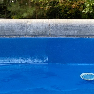 May include: A close-up shot of a blue pool with a concrete edge. The pool's surface is a vibrant blue, with a light fixture visible at the bottom right. The concrete edge is gray and textured, with a crack visible.