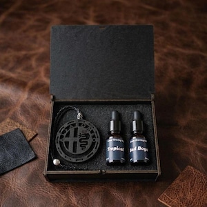 May include: A black wooden gift box containing a circular black pendant with the Alfa Romeo logo on a cord, alongside two small glass dropper bottles labelled Tropical and Bad Boyz. The interior of the box is lined with black foam padding.