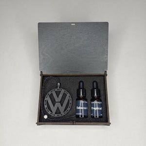 Pode incluir: Uma caixa de madeira escura contendo um pingente circular preto com o logótipo VW recortado num cordão entrançado, juntamente com dois pequenos frascos conta-gotas de vidro âmbar rotulados como Tobacco e Tropical. O interior tem espuma preta.