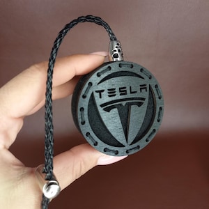 May include: A circular black wooden keychain featuring the Tesla logo and brand name engraved on the front. The piece includes a braided black cord attached to a silver-toned metal connector at the top and a silver-toned cylindrical bead at the base.