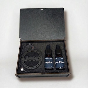 May include: A black wooden gift box containing a circular black pendant with the word Jeep engraved in the centre, attached to a black braided cord. Next to the pendant are two small glass dropper bottles with labels reading Tropical and Bad Boys.