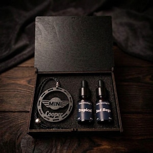 Peut inclure: Une boîte en bois sombre contenant un pendentif circulaire noir avec le texte MINI Cooper et un logo ailé, accompagné de deux petits flacons compte-gouttes en verre avec des étiquettes Laser Graving Oil, Tropical et Bad Boys.