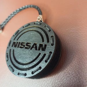 Puede incluir: Un llavero circular de madera negra con el logotipo de Nissan grabado en el centro. El disco tiene un detalle de borde cosido y está unido a un cordón trenzado negro fino con un conector metálico plateado en la parte superior de la pieza.