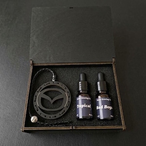 Puede incluir: Una caja de regalo de madera negra que contiene un colgante circular negro con el logotipo de Mazda en un cordón trenzado y dos frascos cuentagotas de vidrio. Las etiquetas tienen el texto Lazer Graving UK, Tropical y Bad Boys en blanco.