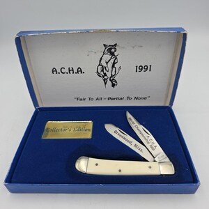 May include: A collector's edition pocket knife in a blue box. The knife has a cream-colored handle and two blades, one engraved with "Greenwood, Miss." and "World Championship 1991 A.C.H.A.". The box lid features a raccoon illustration and the text "A.C.H.A. 1991".