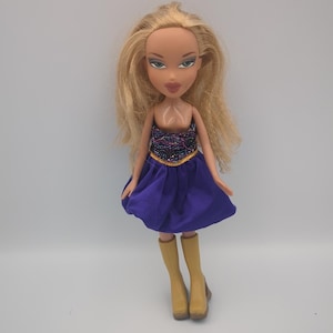 May include: A Bratz doll with long blonde hair, wearing a purple skirt, a sequined top with gold trim, and tan boots. The doll has makeup and is posed against a plain background. The doll is a collectible toy.