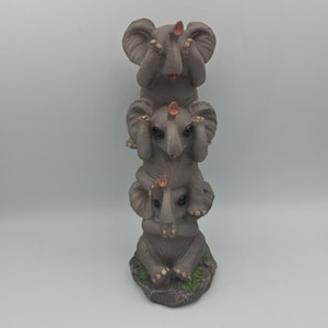 May include: A grey figurine of three elephants stacked. Each elephant covers its eyes with its trunk. The elephants are on a stone base with green detailing.