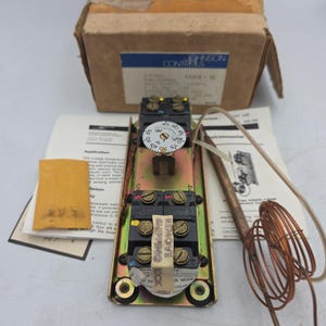 May include: A Johnson Controls A35A18-50 temperature control unit with a circular white dial marked 65 to 95 degrees Fahrenheit, black terminal blocks with brass screws, a copper sensing coil, instruction manuals, and a cardboard box.