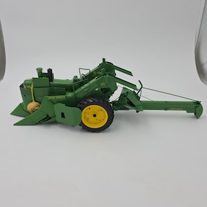 May include: A green and yellow John Deere combine model. The model features a detailed design with a large front wheel, a cab, and various mechanical components. The John Deere logo is visible on the side. The model is set against a white background.
