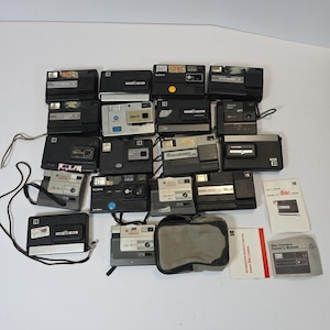 May include: A collection of vintage black and silver film cameras, including Kodak and other brands. Some cameras have attached straps. Also included are a gray camera case and instruction manuals.