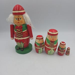 May include: A set of four wooden nesting dolls painted in red, green, and gold. The largest doll is a nutcracker figure with white faux hair, a red hat, and a green base. The three smaller dolls feature matching red and green outfits with painted faces.