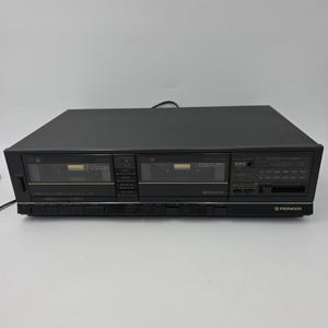 May include: A black Pioneer dual cassette deck featuring two cassette compartments, a central control panel with various buttons, and a digital display area on the right side. The unit has a rectangular shape with a matte finish and labeled controls.