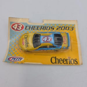 Puede incluir: Un coche de carreras de juguete amarillo y azul, empaquetado en una tarjeta amarilla. El coche tiene el número 43 y el logotipo de Cheerios. El embalaje dice "Cheerios 2003".