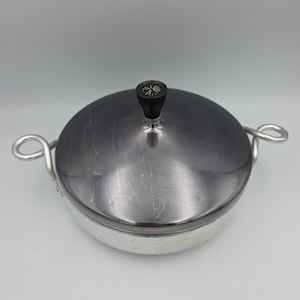 May include: A round metal cooking pot with a brushed silver finish and two looped side handles. The pot features a dark, polished metal lid with a black knob handle on top, which is decorated with a small, gold-colored starburst graphic design.