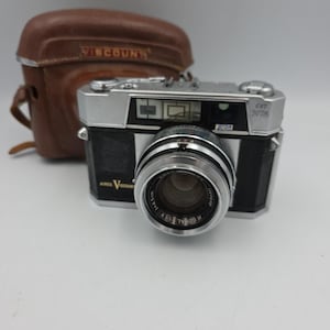 May include: Vintage silver and black camera with a large lens, accompanied by a brown leather case. The camera has the text "AIRES" and "VISCOUNT" on it. The lens has the text "H CORAL" and "1:1.9 f=4.5cm".