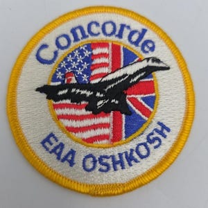 Può includere: Una toppa circolare con bordo giallo. Il testo "Concorde" in blu è sopra un disegno che combina le bandiere americana e britannica. Una silhouette di aereo in bianco e nero è centrata sulle bandiere. Il testo "EAA OSHKOSH" è sotto.