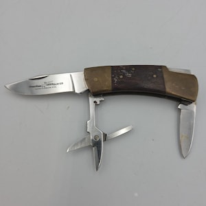 May include: A multi-tool pocket knife with a stainless steel blade and a wooden handle. The knife has a small blade, scissors, and a screwdriver. The text "DEERSLAYER" is visible on the blade.