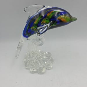 May include: A glass dolphin figurine featuring a swirling pattern of deep blue, green, and orange colors. The dolphin is positioned in a leaping pose, supported by a clear, textured glass base that resembles splashing water.