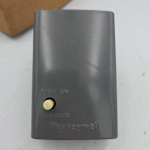 May include: A rectangular gray Honeywell control switch featuring a small cream-colored circular button. The text PUSH ON is printed above the button, and PULL AUTO is printed below it. The device has a smooth, solid plastic casing.