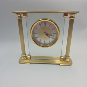 May include: A gold-toned quartz clock with a round face and Roman numerals. The clock is encased in a rectangular frame with two vertical columns and a glass panel. The clock face reads "Baden Park".
