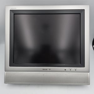 May include: A silver Sharp Aquos television with a black screen frame. The device features a perforated speaker grille along the bottom edge, with the brand name Sharp centered and the word Aquos in the top left corner. Power and sleep indicators are visible.