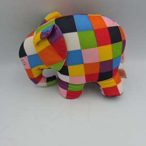 May include: A plush elephant toy featuring a vibrant, multi-colored patchwork pattern of squares in shades of red, yellow, blue, green, black, and pink. The stuffed animal has a trunk, large ears, and a soft fabric texture throughout its body.