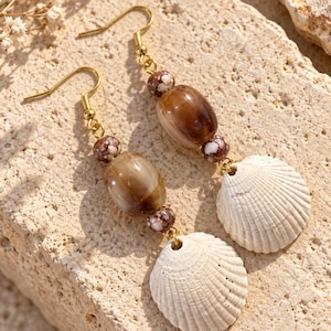 May include: A pair of dangle earrings featuring gold-toned hooks, brown marbled oval beads, small round brown and white speckled beads, and white ribbed seashell pendants. The earrings are arranged vertically with the beads and shells suspended below.