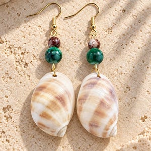 May include: A pair of drop earrings featuring natural cowrie-style shells suspended from gold-toned hooks. Each earring includes a small, round, mottled brown bead and a larger, smooth, vibrant green bead positioned above the shell pendant.