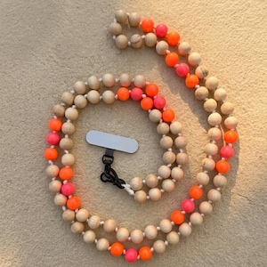 May include: A wooden beaded phone strap with alternating orange and pink beads. The strap has a black clasp and a white connector for attaching to a phone. The beads are strung on a white cord.