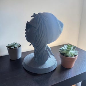 Puede incluir: Un busto escultórico de color gris oscuro que representa una cabeza con rastas, un gorro con un logotipo circular y gafas de sol. La figura se asienta sobre una base circular, capturada de perfil, mostrando los detalles texturizados del cabello.