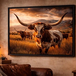 May include: A framed canvas print featuring a herd of Texas Longhorn cattle in a field of tall grass. The cattle are brown and white with large horns. The background shows a sunset with mountains and a cloudy sky.