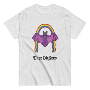 May include: A light gray t-shirt featuring a purple bat with a rainbow and the text "Thee Oh Sees". The bat has outstretched wings and is centered above the text. The rainbow has multiple colors.