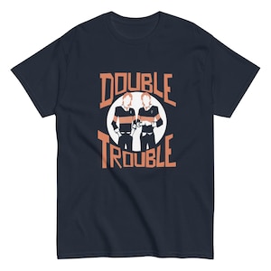 以下が含まれることがあります： オレンジ色の「DOUBLE TROUBLE」の文字と、黒とオレンジのストライプのシャツを着た2人の人物のグラフィックが描かれた、ネイビーブルーのTシャツ。白い円の中に配置されています。