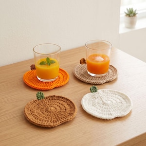 Könnte beinhalten: Vier runde, gehäkelte Untersetzer in Kürbisform, jeweils mit einem kleinen grünen Stiel an der Oberseite. Die Untersetzer sind orange, beige, braun und weiß. Zwei klare Gläser mit Orangensaft stehen auf den orangefarbenen und beigen Untersetzern.