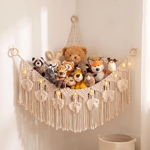 May include: A cream-colored macrame corner hammock for stuffed animals, featuring a triangular woven net design with decorative fringe tassels and wooden rings. The hammock is filled with various plush toys, including a bear, fox, and zebra.