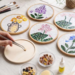 Puede incluir: Un conjunto de posavasos circulares de madera con diseños florales y botánicos de mosaico hechos de pequeñas piezas de vidrio. Una base vacía se trabaja con pinzas metálicas, junto a cuencos con piezas sueltas, pegamento y pinceles.