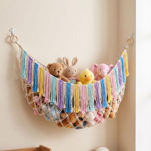 Könnte beinhalten: Eine cremefarbene Spielzeughängematte aus Makramee mit dekorativen Fransen in Pastelltönen wie Blau, Lila, Rosa und Gelb. Das Netz ist an zwei weißen Wandhaken befestigt und enthält mehrere Plüschtiere, darunter einen Bären, einen Hasen und eine Ente.
