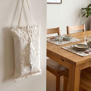Peut inclure: Un porte-mouchoirs en tissu crème orné de délicates broderies florales blanches avec une ouverture verticale centrale. La pochette rectangulaire comprend une sangle en tissu assortie pour la suspension, avec un mouchoir blanc visible.