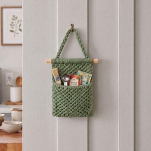 Puede incluir: Un bolsillo de macramé para colgar en la pared de color verde salvia con un asa trenzada suspendida de un pasador de madera. El bolsillo contiene un pequeño cepillo de madera, una cuchara medidora plateada y dos sobres de especias.
