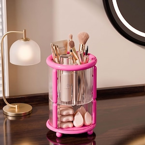 May include: A cylindrical rotating makeup organizer with a bright pink frame and clear, vertically ribbed acrylic panels. The two-tier storage unit holds various cosmetic brushes, a tube labeled Armani Code, a bottle labeled Cia, and makeup sponges.