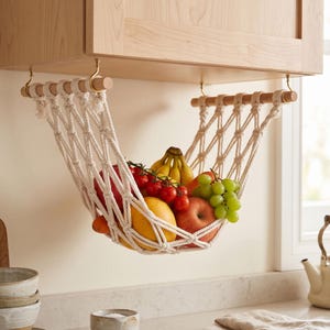 Peut inclure: Un hamac à fruits en macramé fait de corde de coton blanc cassé, suspendu entre deux tiges en bois clair avec des crochets en laiton. Le hamac contient divers produits frais, dont une grappe de bananes, des tomates rouges, une mangue, une pomme et du raisin vert.