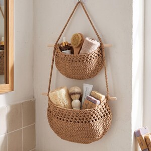 May include: A two-tiered hanging storage organiser featuring two tan crocheted baskets suspended by braided cords from light wooden dowels. The baskets contain various bathroom items including a brush, a folded cloth, soap, and a tube of toothpaste.