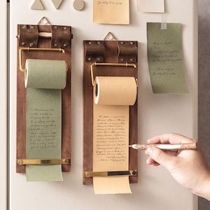May include: Two vertical wooden wall-mounted note dispensers with brass hardware and leather accents. Each holds a roll of paper, one sage green and one tan, featuring handwritten cursive text. A textured wooden pen is held near the tan paper roll.