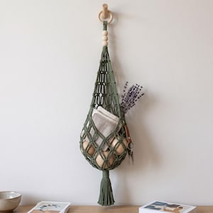 May include: A sage green macrame hanging storage basket featuring a knotted net design, a wooden ring at the top, three light wooden beads, and a bottom tassel. The basket holds several light brown eggs, a small white pouch, and dried lavender.