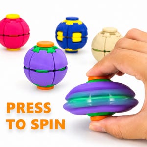 Op de afbeelding: Een verzameling bolvormige fidget-speeltjes in roze, blauw, beige en paars met oranje of gele accenten. Eén paars en groen speeltje wordt getoond in een platgedrukte, draaiende toestand. De tekst PRESS TO SPIN staat in oranje aan de linkerkant.