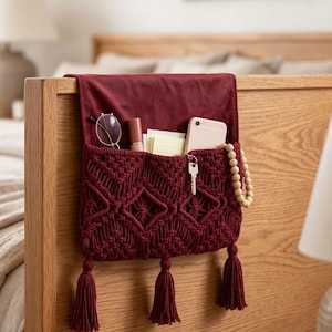 May include: A deep red macrame bedside organiser featuring a flat top flap, a woven diamond pattern, and three hanging tassels at the bottom. The organiser holds a pair of glasses, a lipstick, a notepad, a smartphone, a key, and a wooden bead strand.